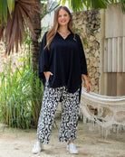 Woman wearing a black top and patterned pants standing outdoors with plants and a hammock in the background.