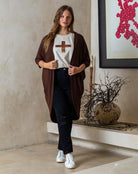 Woman wearing a brown cardigan, white t-shirt with a cross design, and black pants in a room with a decorative vase and artwork.