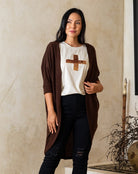 Woman wearing a brown cardigan over a white shirt with a cross design, standing against a beige wall.