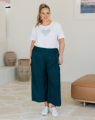 Woman wearing a white t-shirt with a heart design and teal pants in an indoor setting.