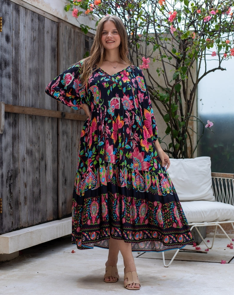 Woman wearing a colorful floral dress standing outdoors with a wooden wall and white chair in the background.