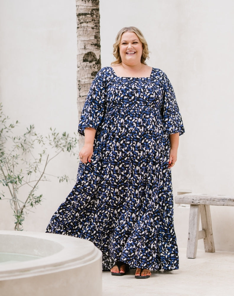 A woman modeling a navy blue maxi dress with a white abstract floral pattern, standing in a bright, modern outdoor setting with a white wall, a bench, and a birch tree trunk.