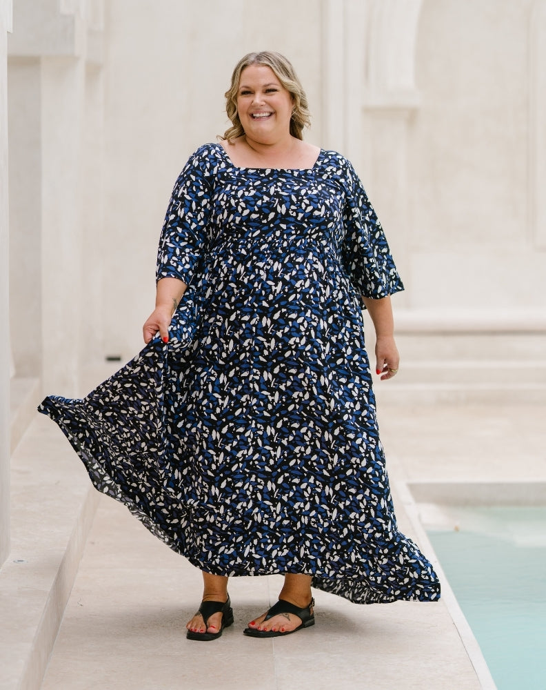 A woman wearing a navy blue and white floral print maxi dress with elbow-length sleeves, walking outdoors next to a light-colored stone structure and a pool.