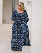 A woman wearing a navy blue maxi dress with a white and light blue abstract floral pattern, standing in a bright, modern outdoor corridor.