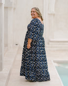 A woman wearing a navy blue maxi dress with a white and light blue abstract floral pattern, standing in a bright, sunlit outdoor patio setting with white columns and a pool visible.
