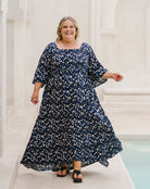 A woman wearing a navy blue and white floral print maxi dress with three-quarter sleeves and a square neckline, walking outdoors in front of a light-colored stone building.