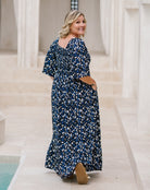 A woman wearing a navy blue maxi dress with a white and light blue abstract floral pattern, standing in a bright, modern courtyard with white columns.
