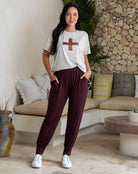 Woman wearing a white t-shirt with a brown design and maroon pants standing in a living room.