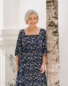 A woman with short gray hair wearing a navy blue dress with a white and light blue abstract pattern, standing next to a tree trunk in front of a white building.