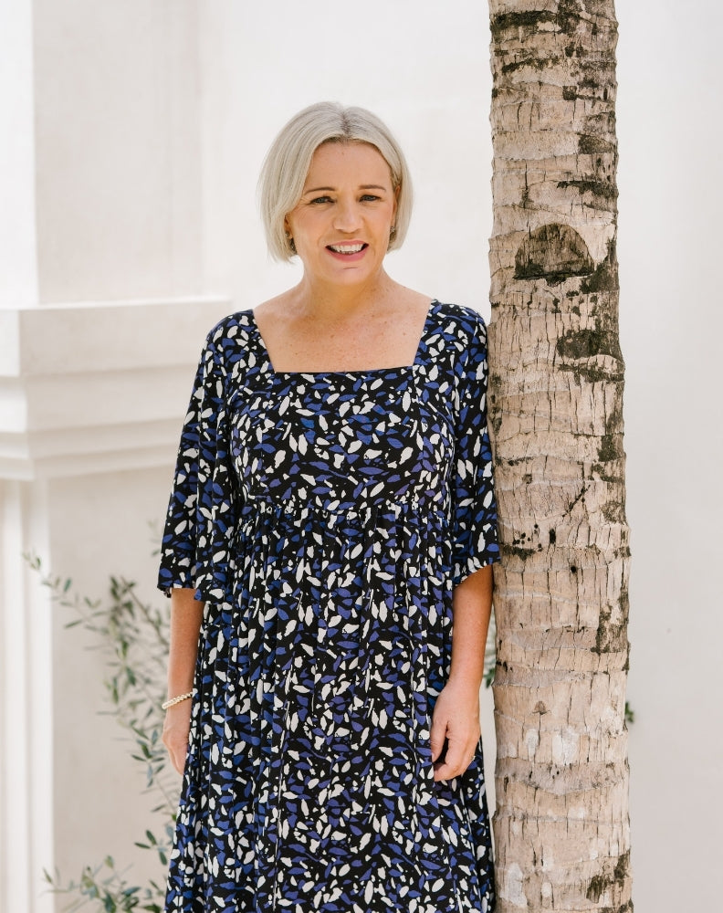 A woman with short gray hair wearing a navy blue dress with a white and light blue abstract pattern, standing next to a tree trunk in front of a white building.