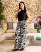 Woman wearing a black sleeveless top and patterned pants standing in a living room with a stone wall background.