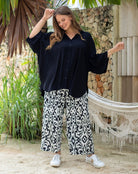 Woman wearing a black shirt and patterned pants standing outdoors with plants and a hammock in the background.