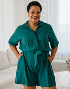Woman wearing a teal romper standing in a bright room with white curtains.