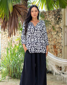 Woman wearing a black and white patterned top and black pants standing in a tropical setting with plants and a stone wall.