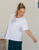 Woman wearing a white t-shirt with a heart design, standing indoors against a plain wall.