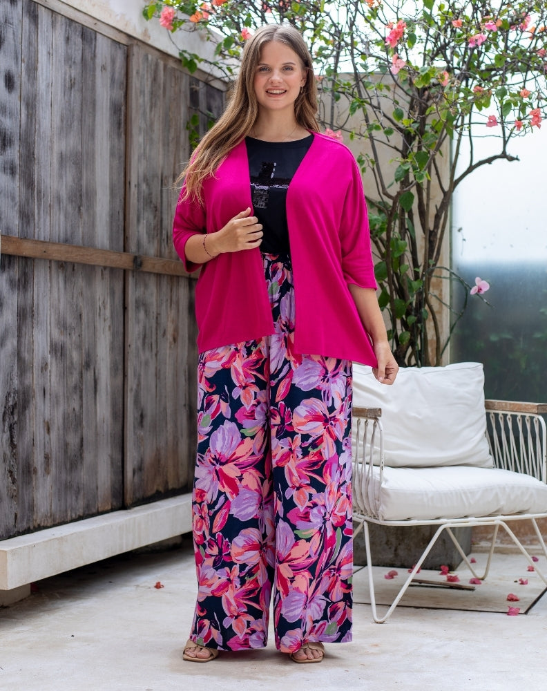 Woman wearing a bright pink cardigan and colorful floral pants standing on a patio.