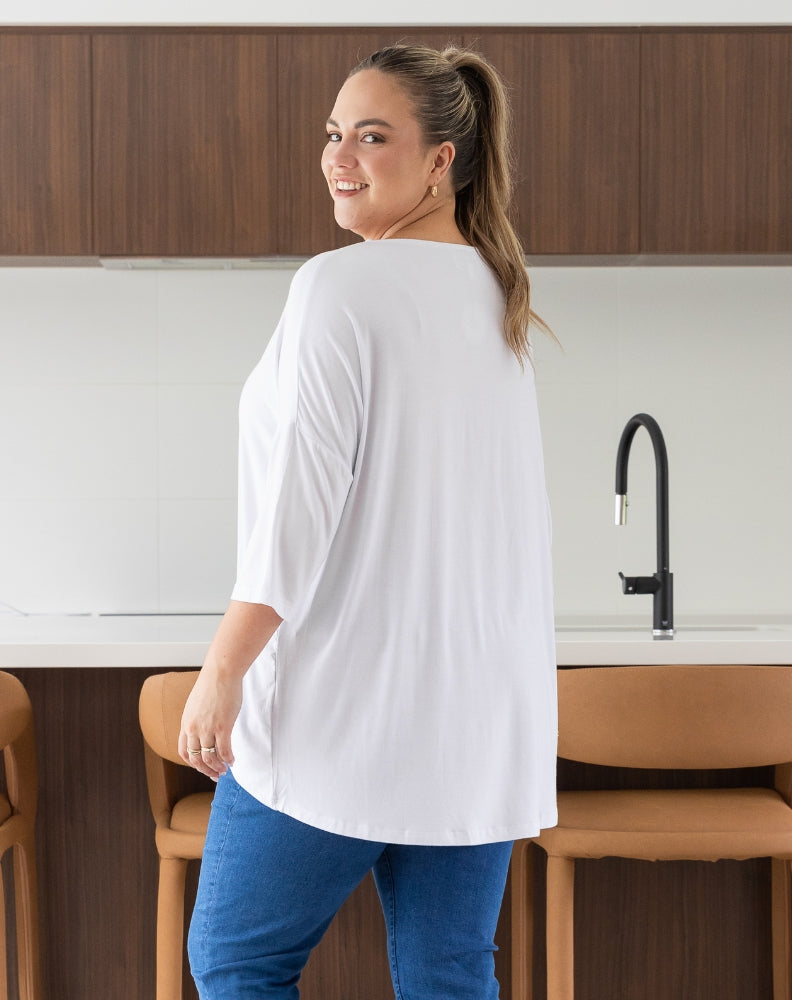 Woman standing in a kitchen wearing a white shirt and blue pants.