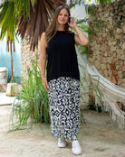 Woman wearing a black sleeveless top and patterned skirt standing outdoors with plants and a hammock in the background.