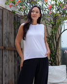 Woman wearing a white sleeveless top and black pants standing outdoors with a wooden fence and plants in the background.