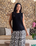 Woman wearing a black sleeveless top and patterned pants standing in a room with a stone wall and decorative elements.