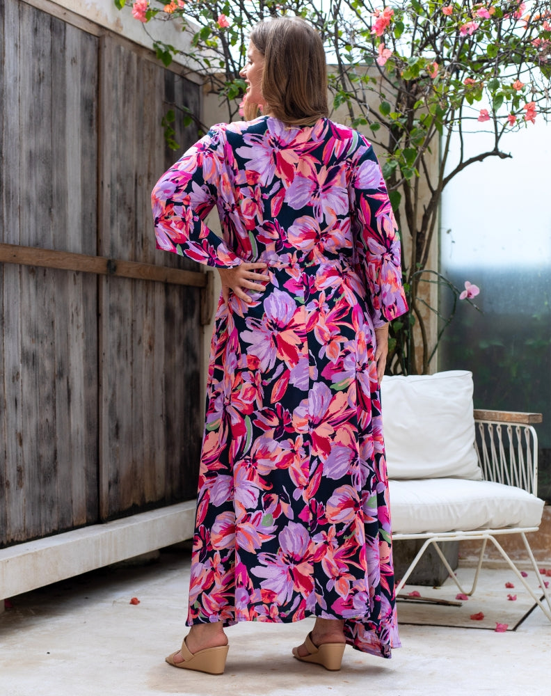 Woman wearing a colorful floral dress standing outdoors near a wooden fence and white chair.