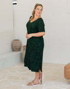 Woman wearing a green patterned dress standing in a room with white walls and a beige floor.