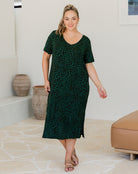 Woman wearing a green patterned dress standing in a room with a white wall and beige floor.
