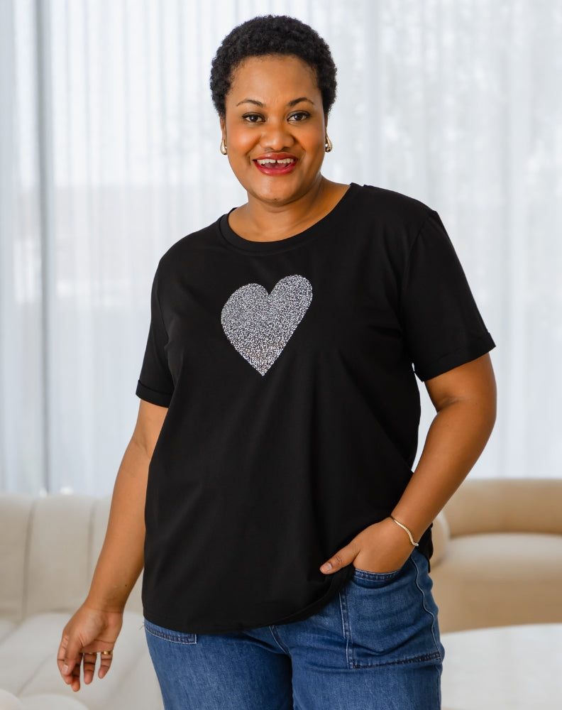 Woman wearing a black t-shirt with a heart design, standing in a room with white curtains.