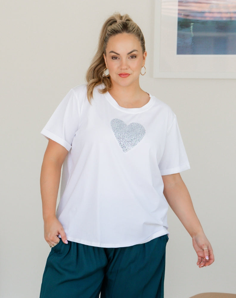 Woman wearing a white t-shirt with a heart design and teal pants indoors.