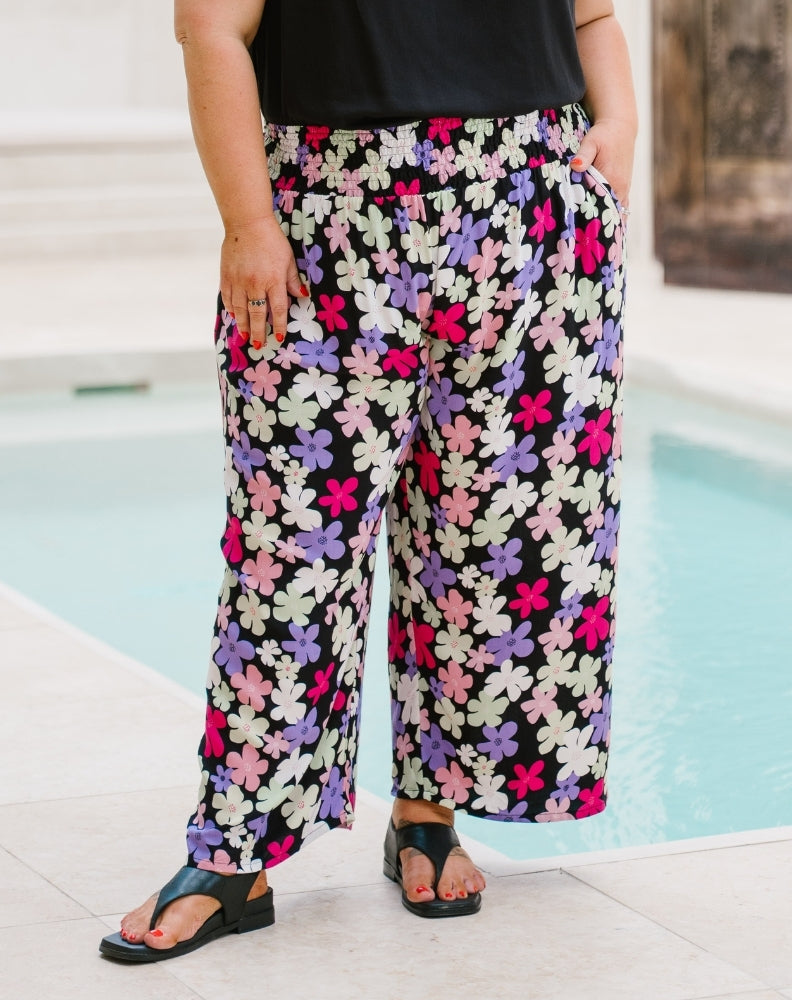 A person wearing black wide-leg cropped pants with a colorful floral print in pink, purple, and white, standing on a patio next to a swimming pool.