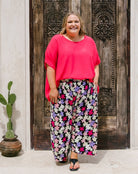 A woman stands in front of a carved dark wooden door wearing a bright pink short-sleeve top and black wide-leg pants with a colorful floral print. A small potted cactus sits on the left.