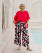 A woman stands outdoors next to a light-colored pool, wearing a bright red short-sleeve top and high-waisted, wide-leg floral pants with pink, purple, and white flowers on a dark background. She is wearing pink-and-white checkered slip-on shoes.