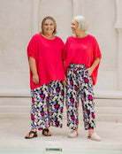 Two women standing side-by-side wearing bright pinkish-red short-sleeve t-shirts and wide-leg floral pants with a black, white, pink, and purple pattern.