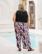 A woman viewed from behind wearing a black sleeveless top and wide-leg pants with a colorful floral print in pink, purple, white, and green, standing by a swimming pool.