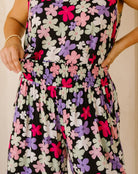 A close-up of a sleeveless jumpsuit with a black background and a colorful floral pattern of pink, purple, and light green flowers. The jumpsuit features a smocked elastic waistband and is modeled by a person whose hands and arms are visible.
