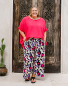 A woman modeling a bright pink short-sleeve top and black wide-leg pants with a colorful floral print, standing in front of a rustic dark wooden door.