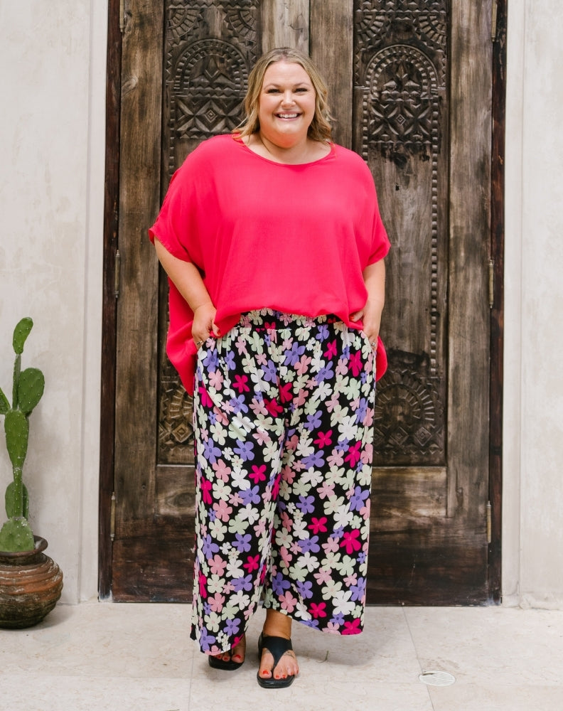 A woman modeling a bright pink short-sleeve top and black wide-leg pants with a colorful floral print, standing in front of a rustic dark wooden door.