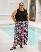 A woman stands by a swimming pool wearing a sleeveless black top and wide-leg floral pants with a purple, pink, and white pattern on a dark background.