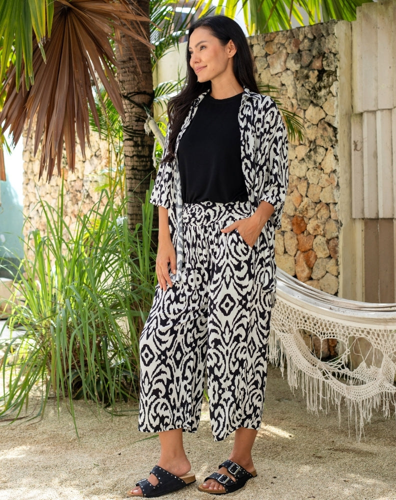Woman in a black and white patterned outfit standing outdoors with plants and a stone wall in the background.