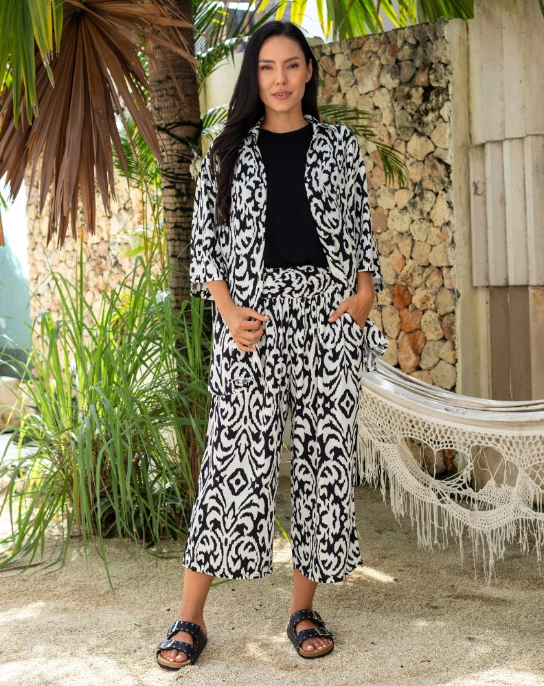 Woman in a black and white patterned outfit standing outdoors with plants and a hammock in the background.