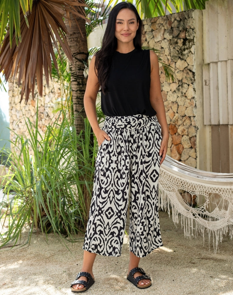 Woman wearing a black sleeveless top and patterned pants standing in a tropical setting with plants and a hammock.