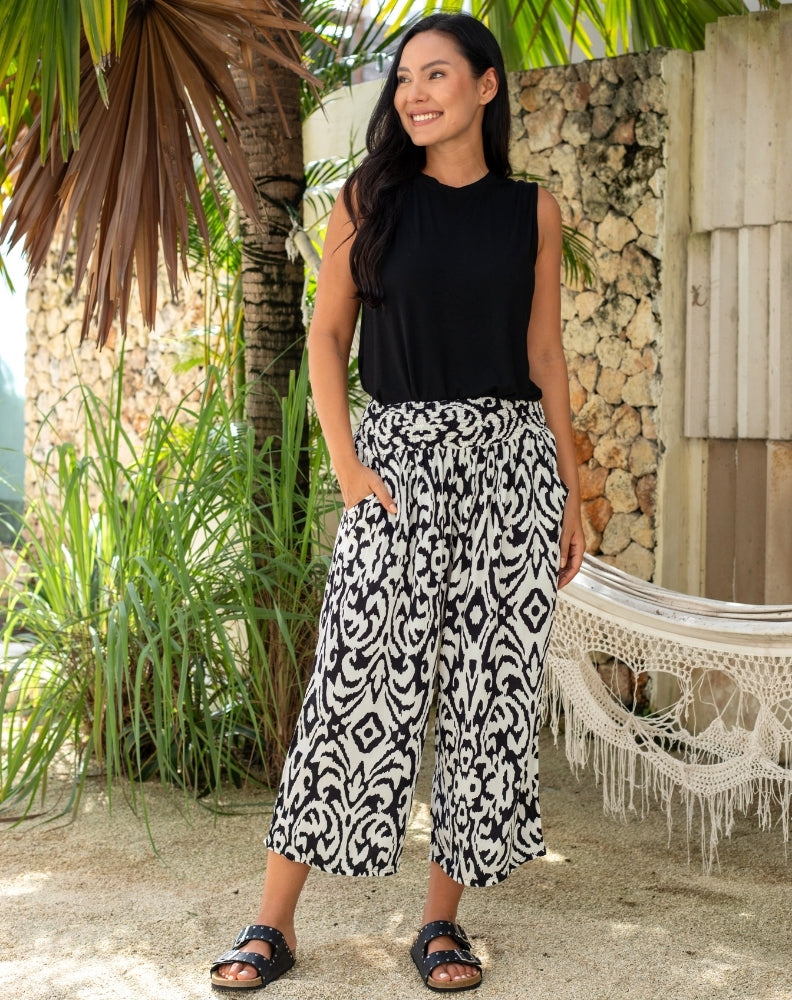 Woman wearing a black sleeveless top and patterned pants standing outdoors with plants and a hammock in the background.