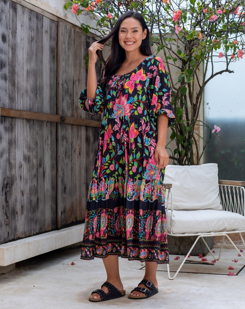 Woman wearing a colorful floral dress standing outdoors with a wooden wall and white chair in the background.