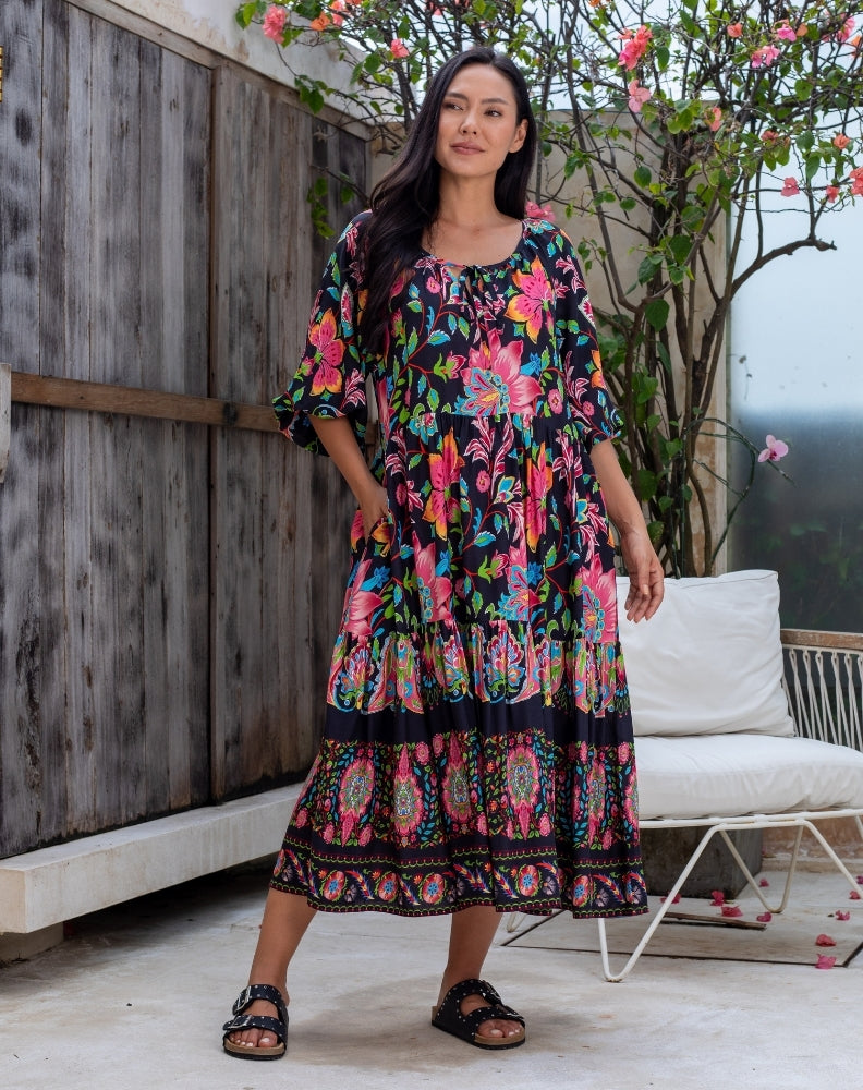 Woman wearing a colorful floral dress standing outdoors with a wooden wall and white chair in the background.