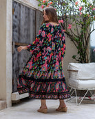 Woman wearing a colorful floral dress standing in an outdoor setting with a wooden door and white chair.