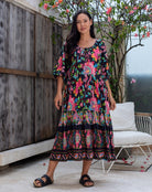 Woman wearing a colorful floral dress standing outdoors with a wooden wall and white chair in the background.