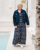 A woman walking outdoors in a navy blue denim jacket over a navy, white, and light blue floral maxi dress, paired with white sneakers.