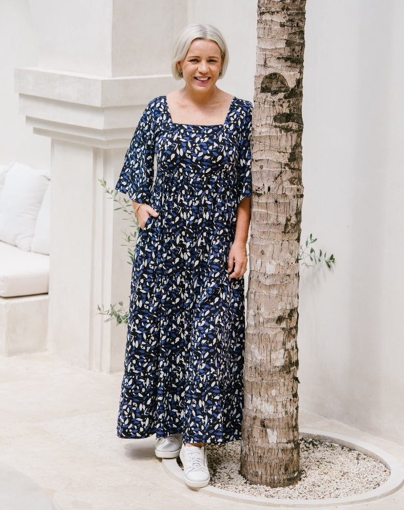 A woman with short gray hair wearing a navy blue and white floral print maxi dress, standing next to a tree trunk in a bright, modern outdoor setting with white walls.