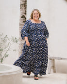 A woman modeling a navy blue maxi dress with a white and light blue abstract floral print, standing in a bright, minimalist outdoor setting with a white wall, a tree trunk, and a rustic bench.