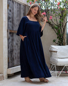 Woman in a navy blue dress standing outdoors with a wooden door and white chair in the background.